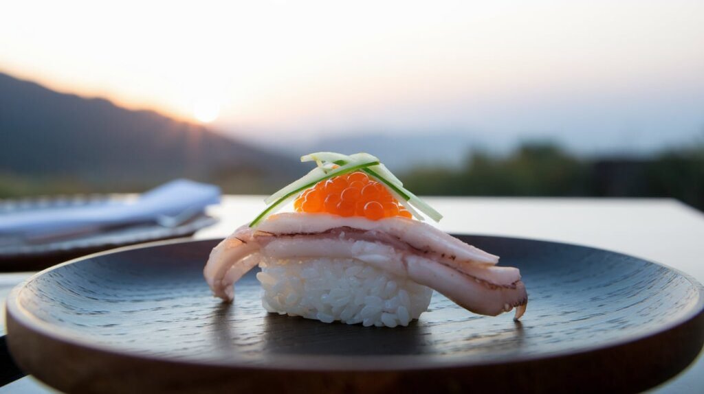 Tako sushi topped with fresh octopus slices, garnished with salmon roe, served on a wooden plate against a serene sunset background, showcasing the elegance of Japanese cuisine.